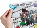 A hand holds a colorful piece of the game 'The Key Murder at the Oakdale' by HABA. The game features intricate artwork with characters behind bars, linked by chains, showing vibrant tokens attached to a clear plastic backdrop.