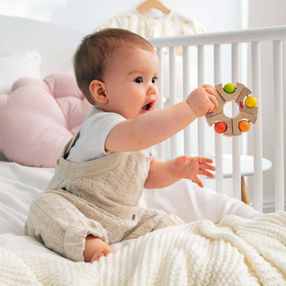 A delighted baby sitting on a soft blanket, reaching out to grasp a colorful wooden grasping toy ball wheel by Haba. The baby wears a cozy light-colored outfit and is surrounded by a serene and soothing nursery environment.