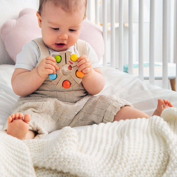 A baby sitting on a bed, holding a colorful Grasping Toy Ball Wheel by Haba. The toy features wooden rings with vibrant colored balls attached. The baby is dressed in a light shirt and beige overalls, looking curious and engaged.