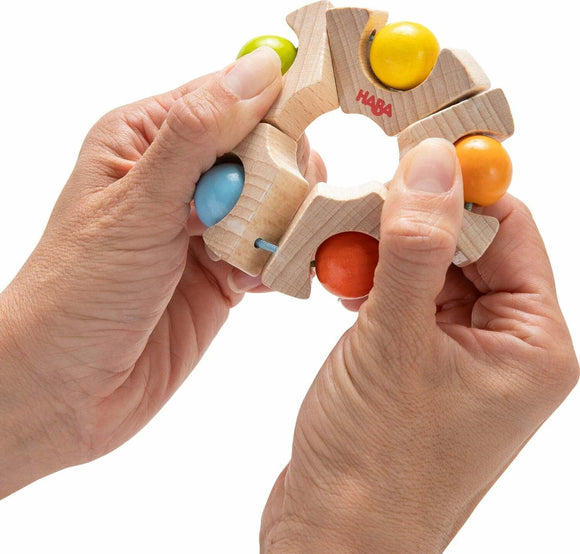 A pair of hands holding the Haba Grasping Toy Ball Wheel. The toy features a circular wooden structure with colorful balls of green, yellow, blue, and orange attached to it. It's designed for infants and toddlers to promote sensory exploration and fine motor skills.