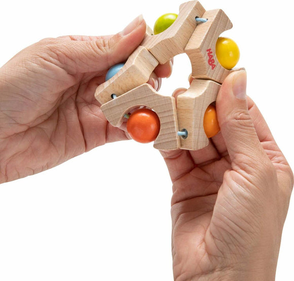 A person holds a colorful Grasping Toy Ball Wheel by Haba. The toy features a circular wooden frame with vibrant balls in blue, green, orange, and yellow, designed for sensory play and motor skill development.