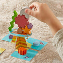 A child’s hand reaching to place a colorful geometric piece onto a stacking game called Flotsam Float, made by Haba. The game features a variety of bright wooden shapes like a star and a gear, resting on a circular base with ocean-themed graphics and surrounded by a sandy surface.
