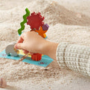 A child’s hand placing a colorful wooden piece onto a sandy surface that features a bright blue game board. The game includes various textured pieces shaped like sea creatures and gears, designed to stimulate creativity and fine motor skills.
