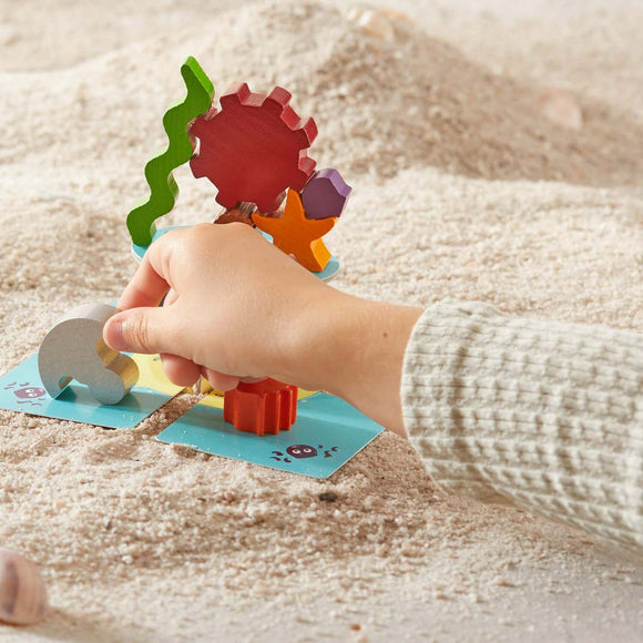 A child’s hand placing a colorful wooden piece onto a sandy surface that features a bright blue game board. The game includes various textured pieces shaped like sea creatures and gears, designed to stimulate creativity and fine motor skills.