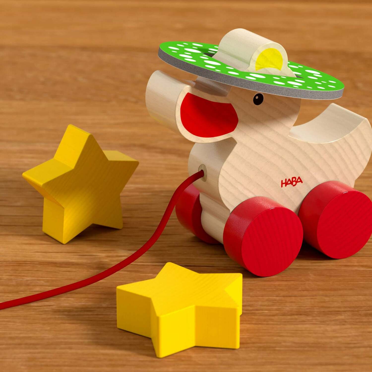 The Duck Game by HABA features a colorful wooden duck toy with a green polka-dotted sombrero and red wheels. Two bright yellow star-shaped pieces are placed nearby, creating an engaging play environment for young children, promoting creativity and motor skills.