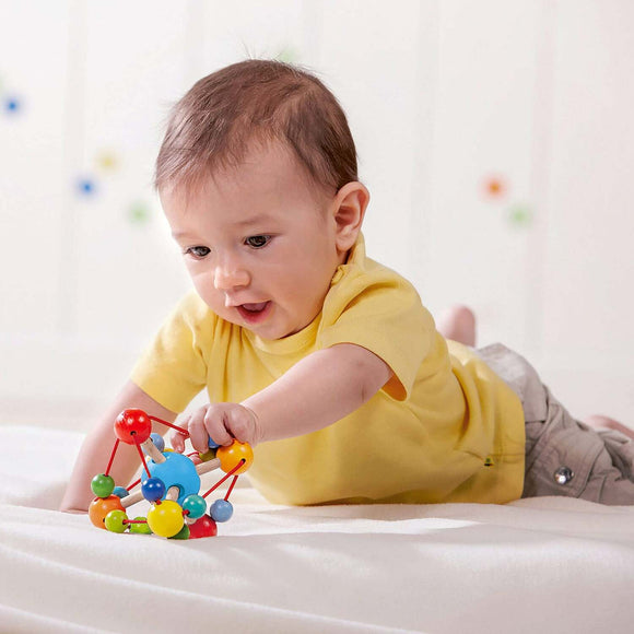 A delighted baby interacting with the Grasping Toy Tirili, made by Haba. The colorful toy features various beads and textures, promoting sensory exploration and motor skill development. The baby, wearing a yellow shirt, is focused and engaged, showcasing the toy's appeal for young children.