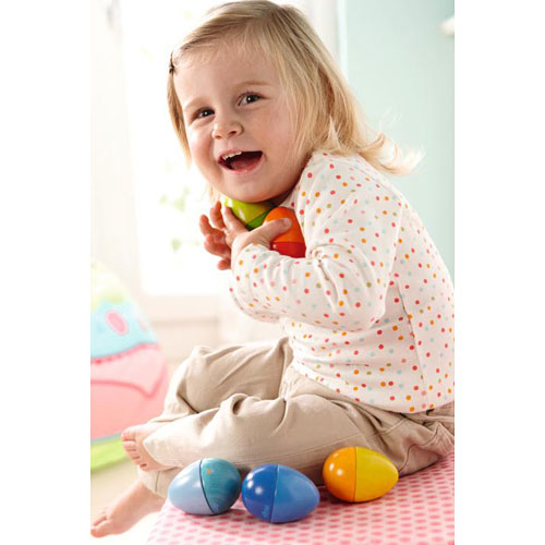 A cheerful child with blonde hair smiles while holding colorful musical eggs. The child is sitting on a soft surface with additional musical eggs nearby, showcasing their playful and engaging design. The background features a bright, cheerful room, enhancing the joyful atmosphere.