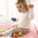 A young child sitting on a light pink cushioned surface plays with colorful musical eggs. The child, wearing a spotted white long-sleeve shirt, holds a blue egg in one hand and an orange one in the other, while several other eggs in different colors, including green and blue, are scattered around.