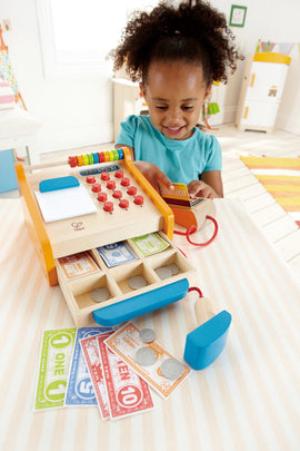 A young girl joyfully playing with a Hape Checkout Register toy. The register features bright colors, buttons, an abacus, and includes play money like bills and coins, promoting imaginative role play and early math skills.