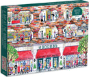 A Day at the Bookstore jigsaw puzzle by Galison featuring a colorful illustration of a bustling bookstore. The scene shows people browsing books, attending a children's storytime, and enjoying a coffee, with detailed storefronts labeled for children's books, history, art, poetry, and more. This 1000-piece puzzle captures the lively atmosphere of a community bookstore.