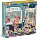 The Book Haven jigsaw puzzle by Galison features a vibrant illustration of a charming bookstore exterior with colorful book displays, a bicycle with a flower basket, and a small dog, showcasing a cozy reading environment. This 1000-piece puzzle invites you to explore the delightful details and whimsical art.