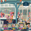 A colorful illustration of an outdoor scene featuring a quaint bookstore titled 'Book Haven.' In the foreground, a vintage bicycle with a woven basket filled with flowers is parked beside the store, which showcases an array of vibrant book covers in its windows. A small dog stands on the patterned sidewalk, completing the charming ambiance of this jigsaw puzzle titled 'Book Haven' by Galison, consisting of 1000 pieces.