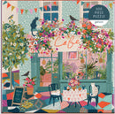 A colorful 500-piece jigsaw puzzle titled 'Afternoon Tea' by Galison. The illustration features a charming café scene adorned with vibrant flowers, playful cats, and decorative bunting. The café, highlighted by a sign reading 'Cafe,' displays a cozy outdoor setting with a round table set for tea, featuring cupcakes and a flower arrangement amidst a bright, patterned floor.