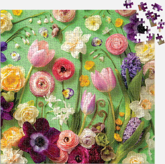 A vibrant 500-piece jigsaw puzzle titled 'Springtime Petals' by Galison featuring an intricate design of colorful flowers including tulips, roses, and daisies on a green background. Some puzzle pieces are scattered around the edge.