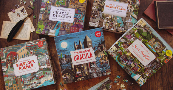 Jigsaw puzzle titled 'The World of Jane Austen' by Laurence King Publishing, featuring colorful illustrations related to Jane Austen's works. The box shows a collage of iconic scenes and characters from her novels. Other puzzle boxes are visible in the background, including titles like 'The World of Sherlock Holmes' and 'The World of Dracula'. A quill and puzzle pieces are scattered on a wooden table.