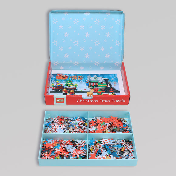 The LEGO Puzzle Christmas Train jigsaw puzzle features a vibrant winter scene with a festive train and colorful LEGO pieces, neatly organized in compartments. The box is decorated with a blue snowflake pattern, adding to the holiday cheer.