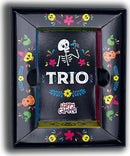 The Trio game by Happy Camper features a colorful box adorned with playful floral designs and a skeleton motif. The box showcases the bold letters 'TRIO' prominently in the center, surrounded by vibrant illustrations of musical instruments, flowers, and whimsical characters, creating a fun and inviting aesthetic for family game nights.