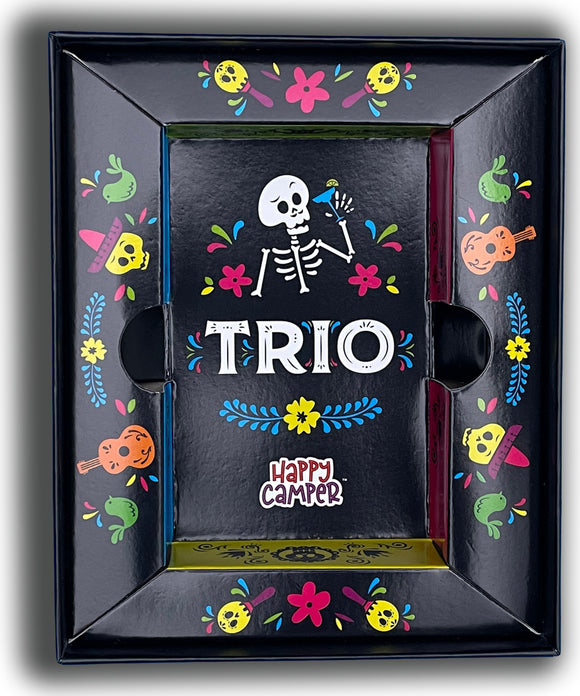 The Trio game by Happy Camper features a colorful box adorned with playful floral designs and a skeleton motif. The box showcases the bold letters 'TRIO' prominently in the center, surrounded by vibrant illustrations of musical instruments, flowers, and whimsical characters, creating a fun and inviting aesthetic for family game nights.