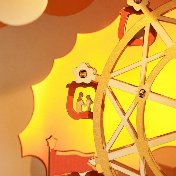 The Music Dreamer Sunset Carnival DIY kit features a beautifully designed Ferris wheel with intricate cutouts depicting a couple inside a cabin. The warm yellow background enhances its playful carnival theme, ideal for craft lovers who enjoy assembling decorative pieces.