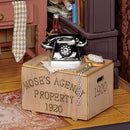 A detailed diorama featuring Mose's Detective Agency. The scene includes a vintage black rotary telephone on a wooden crate labeled 'MOSE'S AGENCY PROPERTY 1920'. Nearby items include a pocket watch, a glass bottle, and scattered papers, all set against a warmly decorated interior with shelves filled with books and a patterned rug on the floor.