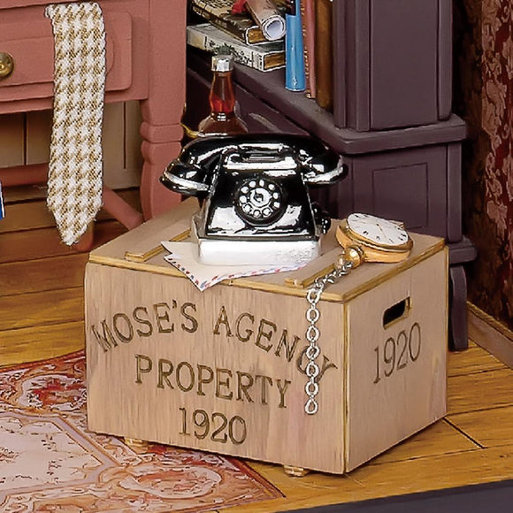 A detailed diorama featuring Mose's Detective Agency. The scene includes a vintage black rotary telephone on a wooden crate labeled 'MOSE'S AGENCY PROPERTY 1920'. Nearby items include a pocket watch, a glass bottle, and scattered papers, all set against a warmly decorated interior with shelves filled with books and a patterned rug on the floor.