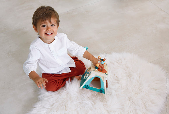 A young boy sits on a soft white rug, joyfully interacting with the Multi Activity Volcano toy by Juratoys. The toy features a vibrant design with different elements for play, including a beaded activity. The boy, dressed in a white shirt and reddish-brown pants, has a cheerful expression as he engages in imaginative play.