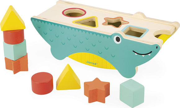 Tropik Shape Sorting Crocodile by Janod, a colorful wooden toy designed for toddlers featuring a crocodile-shaped base with shape sorting holes. The set includes various geometric shapes like circles, squares, and stars in bright colors, promoting fine motor skills and cognitive development through play.