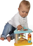 A young child sits on the floor, engaging with the Tropik Multi-Activity Triangle by Janod. The colorful activity triangle features several interactive elements, including spinning gears, a clear spiral tube with colorful beads inside, and stacking blocks decorated with animal prints. The child has a focused expression, exploring the various textures and sounds of the toy, promoting sensory development and fine motor skills.