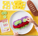 A colorful play set of a 'Math Sandwich' game by MindWare, featuring felt pieces that resemble sandwich ingredients like lettuce, tomato, cucumber, and cheese. A child’s hand is seen arranging the ingredients on a plate. Nearby, there's a checklist card titled '#12 Submarine' that is part of the game, showing different sandwich combinations and a vibrant yellow placemat.