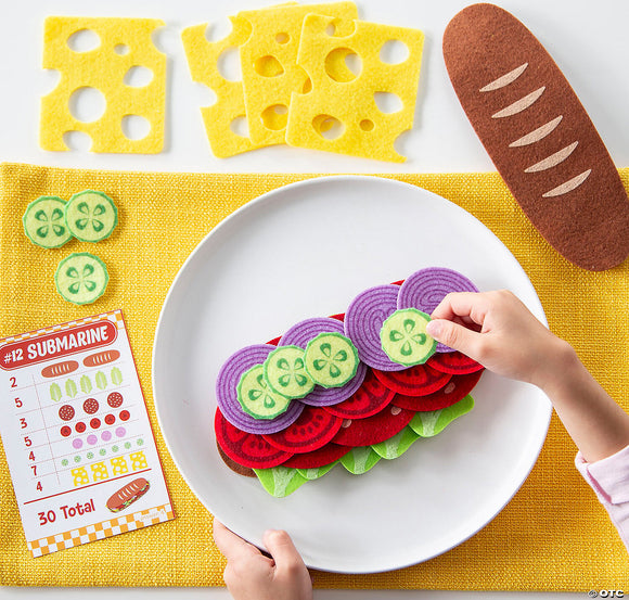 A colorful play set of a 'Math Sandwich' game by MindWare, featuring felt pieces that resemble sandwich ingredients like lettuce, tomato, cucumber, and cheese. A child’s hand is seen arranging the ingredients on a plate. Nearby, there's a checklist card titled '#12 Submarine' that is part of the game, showing different sandwich combinations and a vibrant yellow placemat.