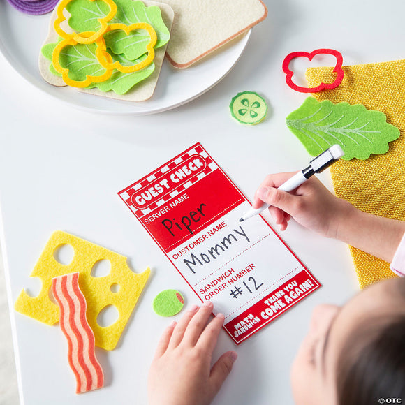 A child is engaged in imaginative play with the Math Sandwich game by MindWare. The scene features colorful felt food items like slices of bread, cheese, lettuce, and bacon, alongside a handwritten guest check listing 'Piper' as the server and 'Mommy' as the customer. This vivid setup encourages creativity and learning through pretend play.