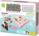 The Busy Busy Bake Shop game features a colorful game box displaying a fun 4-in-a-row donut game for kids. The image shows various wooden donuts in different colors and flavors on a pink checkered game board, along with tongs and dice. Two children are seen playing the game together, emphasizing cooperative play and fine motor skills development.