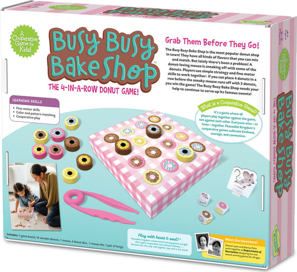 The Busy Busy Bake Shop game features a colorful game box displaying a fun 4-in-a-row donut game for kids. The image shows various wooden donuts in different colors and flavors on a pink checkered game board, along with tongs and dice. Two children are seen playing the game together, emphasizing cooperative play and fine motor skills development.