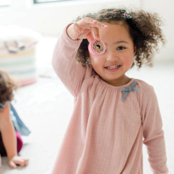A young girl with curly hair smiles brightly while holding up a pink donut toy, showcasing her excitement. She is wearing a pastel-colored dress and standing on a soft floor with another child playing in the background.