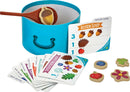 Acorn Soup game by Peacable Kingdom featuring a vibrant blue pot, wooden spoon, and various colorful recipe cards. The game includes charming food tokens like acorns, flowers, and fruits for engaging gameplay. Ideal for interactive playtime, encouraging creativity and social skills.