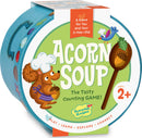 A circular game box for 'Acorn Soup' by Peaceable Kingdom. The box features a friendly cartoon squirrel wearing a chef's hat, holding an acorn. The title 'Acorn Soup' is prominently displayed in bold green letters on a pastel green background. The design is colorful and playful, emphasizing that it's a counting game suitable for children aged 2 and above. It promotes play, learning, and exploration.