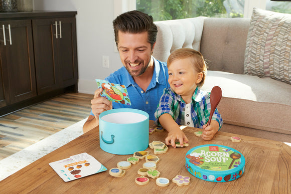 A father and son engage in a fun game of Acorn Soup by Peaceable Kingdom. They are smiling, with the son holding a wooden spoon and looking at game pieces spread across the table. The colorful game components include a round box and illustrated cards, creating an interactive and joyful playtime experience.