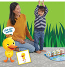 A joyful scene featuring a young child and an adult playing the game 'DUCK DUCK DANCE!' from Peaceable Kingdom. The child is excitedly raising their arms, while the adult is laughing and encouraging them. In the foreground, there is a cute yellow duck character and a game board with colorful cubes, inviting interactive play in a vibrant green setting.