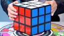 A close-up of a person holding a Rubik's Cube, showcasing the vibrant colored stickers of red, blue, and white, on a game board background. The cube is positioned prominently, emphasizing its intricate design, perfect for magic tricks and puzzles.