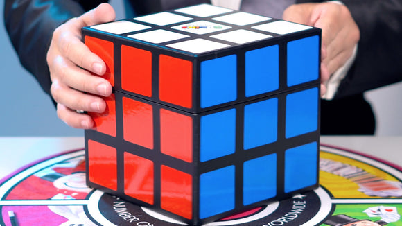 A close-up of a person holding a Rubik's Cube, showcasing the vibrant colored stickers of red, blue, and white, on a game board background. The cube is positioned prominently, emphasizing its intricate design, perfect for magic tricks and puzzles.