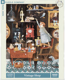 A colorful jigsaw puzzle titled 'Vintage Shop' by New York Puzzle Company featuring a whimsical scene of a vintage store filled with eclectic items. The illustration showcases a cozy shop with a vintage clock, decorative vases, a sewing machine, and various trinkets on shelves. A woman reaches for a bird perched nearby, surrounded by a charming interior with patterned tiles and vintage furniture. This puzzle consists of 1000 pieces.