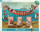 A colorful jigsaw puzzle titled 'All Paws on Deck' featuring whimsical artwork of cats lounging on deck chairs by the seaside, with a woman relaxing on a hammock. A ship is sailing in the background. The puzzle contains 500 pieces and is made by New York Puzzle Company, showcasing a playful and fun beach theme.