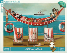 A colorful jigsaw puzzle titled 'All Paws on Deck' featuring whimsical artwork of cats lounging on deck chairs by the seaside, with a woman relaxing on a hammock. A ship is sailing in the background. The puzzle contains 500 pieces and is made by New York Puzzle Company, showcasing a playful and fun beach theme.