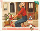 A colorful jigsaw puzzle featuring a woman in a red polka-dot shirt riding a vintage bicycle adorned with a bouquet of flowers. She has a scarf tied around her head and sunglasses on. In the background, there are brownstone buildings with a warm and inviting atmosphere. The puzzle is by New York Puzzle Company and comprises 1000 pieces.