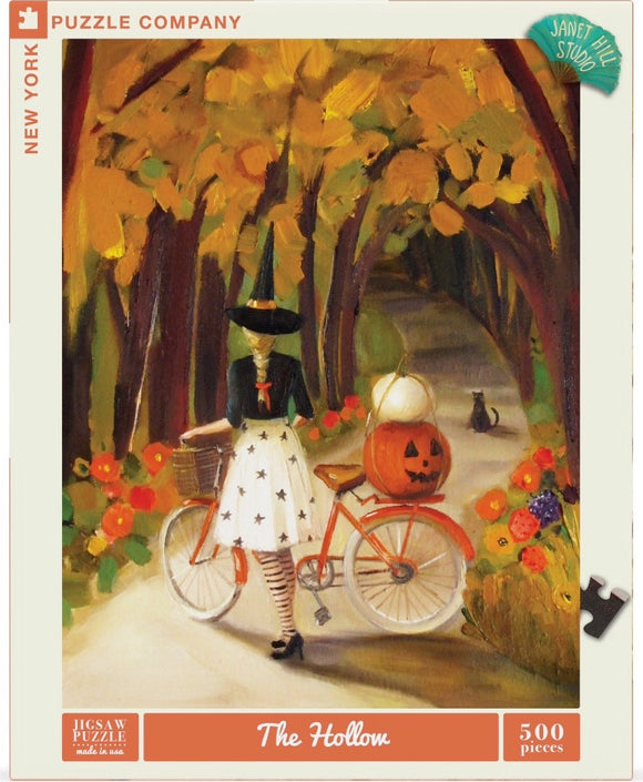 A whimsical jigsaw puzzle titled 'The Hollow' featuring a witch riding a vintage bicycle adorned with a pumpkin. The scene showcases a vibrant autumn forest with colorful leaves and flowers, evoking a charming Halloween atmosphere. The puzzle is designed by Janet Hill Studio and includes 500 pieces.