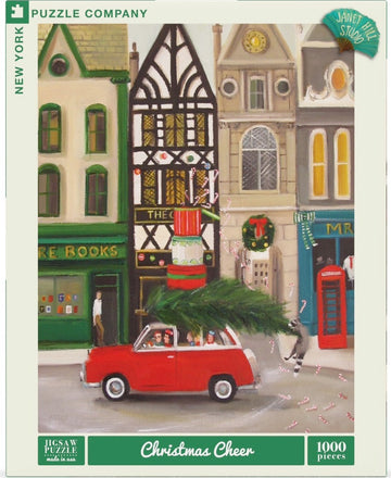 A colorful jigsaw puzzle titled 'Christmas Cheer' by New York Puzzle Company, featuring a festive scene with a red car carrying a Christmas tree, surrounded by charming buildings decorated for the holidays. The puzzle is designed by Janet Hill Studio and includes 1000 pieces.