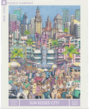 An artistic jigsaw puzzle titled 'Sun Kissed City' by New York Puzzle Company featuring a vibrant scene of Miami Beach. The illustration includes colorful buildings, palm trees, and people enjoying various activities such as dancing and playing music. It showcases a lively beach atmosphere with iconic landmarks and vintage cars, capturing the essence of a sun-drenched city life. The puzzle consists of 500 pieces and is designed for puzzle enthusiasts who appreciate detailed art.