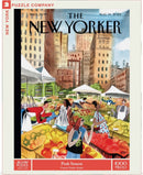 A colorful jigsaw puzzle titled 'Peak Season' by New York Puzzle Company featuring an illustration by Victoria Tentler-Krylov. The design showcases a vibrant farmers' market scene with people engaging in transactions, surrounded by a variety of fresh fruits and vegetables, set against a backdrop of city buildings.