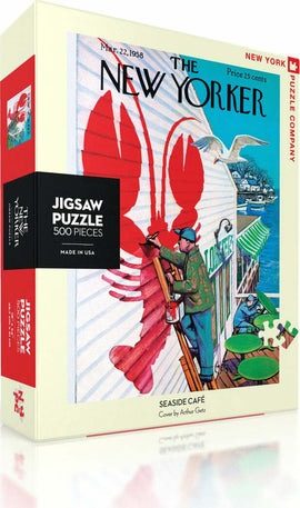 Seaside Cafe jigsaw puzzle by New York Puzzle Company featuring 500 pieces. The box displays a vibrant cover illustration by Arthur Getz, showcasing a picturesque seaside setting with a lobster motif and people engaged in leisurely activities. Perfect for puzzle enthusiasts and art lovers alike.