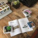 A creative game setup featuring the OuiSi Original: Games of Visua. A colorful stack of cards is placed alongside an open guidebook titled 'Guide to Creative Play' on a wooden table. The background includes a succulent plant and a vintage typewriter.